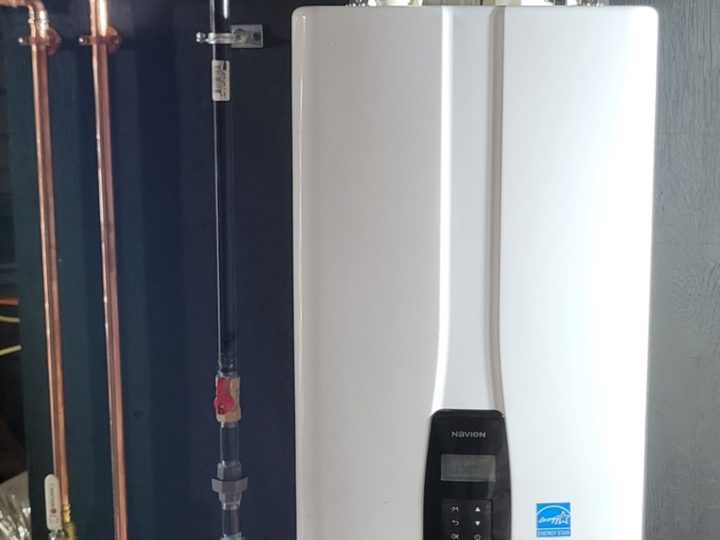 A Navien tankless water heater Dan installed on a dark backer board, with copper supply loops, a gas shutoff and white PVC venting